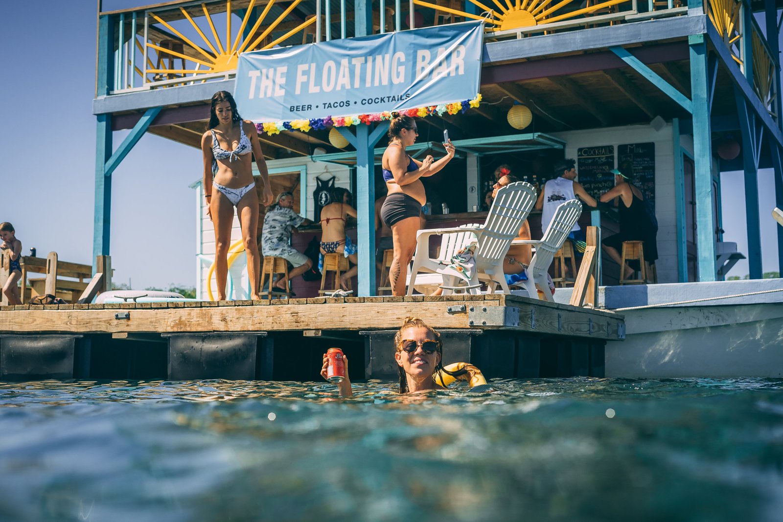 Guests on the swim deck and in the water with a drink below The Floating Bar banner