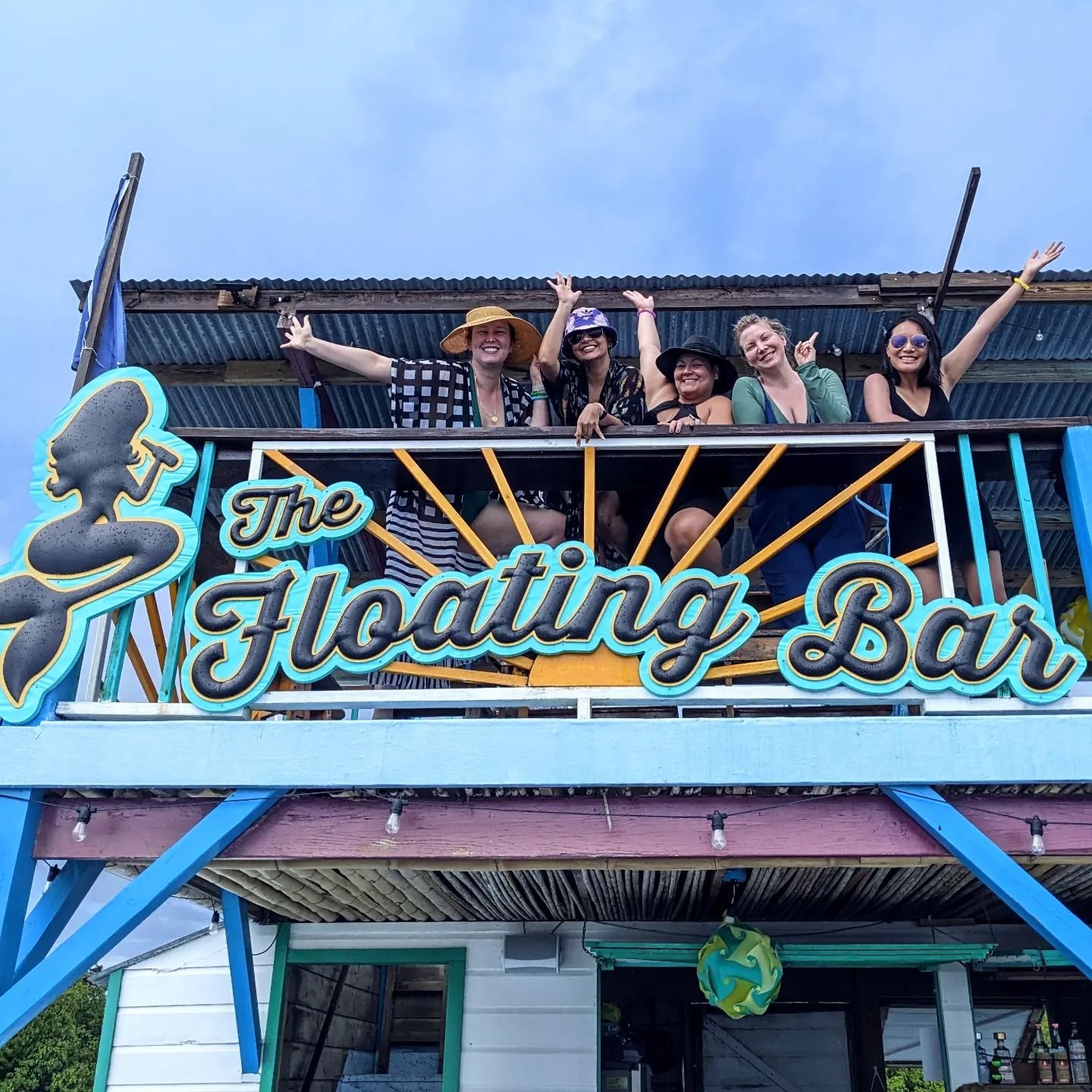 Friends waving from the top deck behind The Floating Bar's mermaid sign